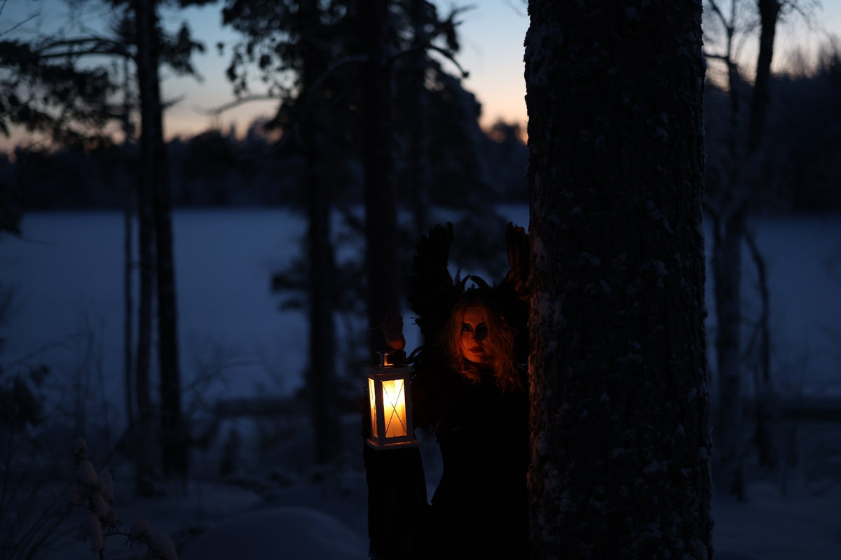 Lyhtyä kantava hahmo puun takana lumisessa metsässä hämärän aikaan.
