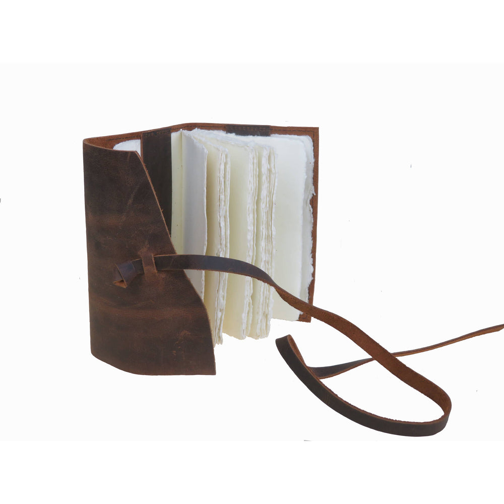 Ruskea nahkakantinen muistikirja raollaan, käsintehdyt paperisivut ja suljentanauha näkyvissä. / Partly open brown leather notebook showing handmade paper pages and a leather tie.