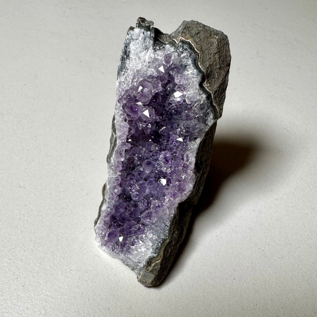 Ametistiklusteri oikealta kuvattuna; kiteiden syvyys ja peruskiven vihreänharmaat sävyt korostuvat. -  Amethyst cluster photographed from the right; the depth of the crystals and the greenish-grey tones of the base rock are emphasized.