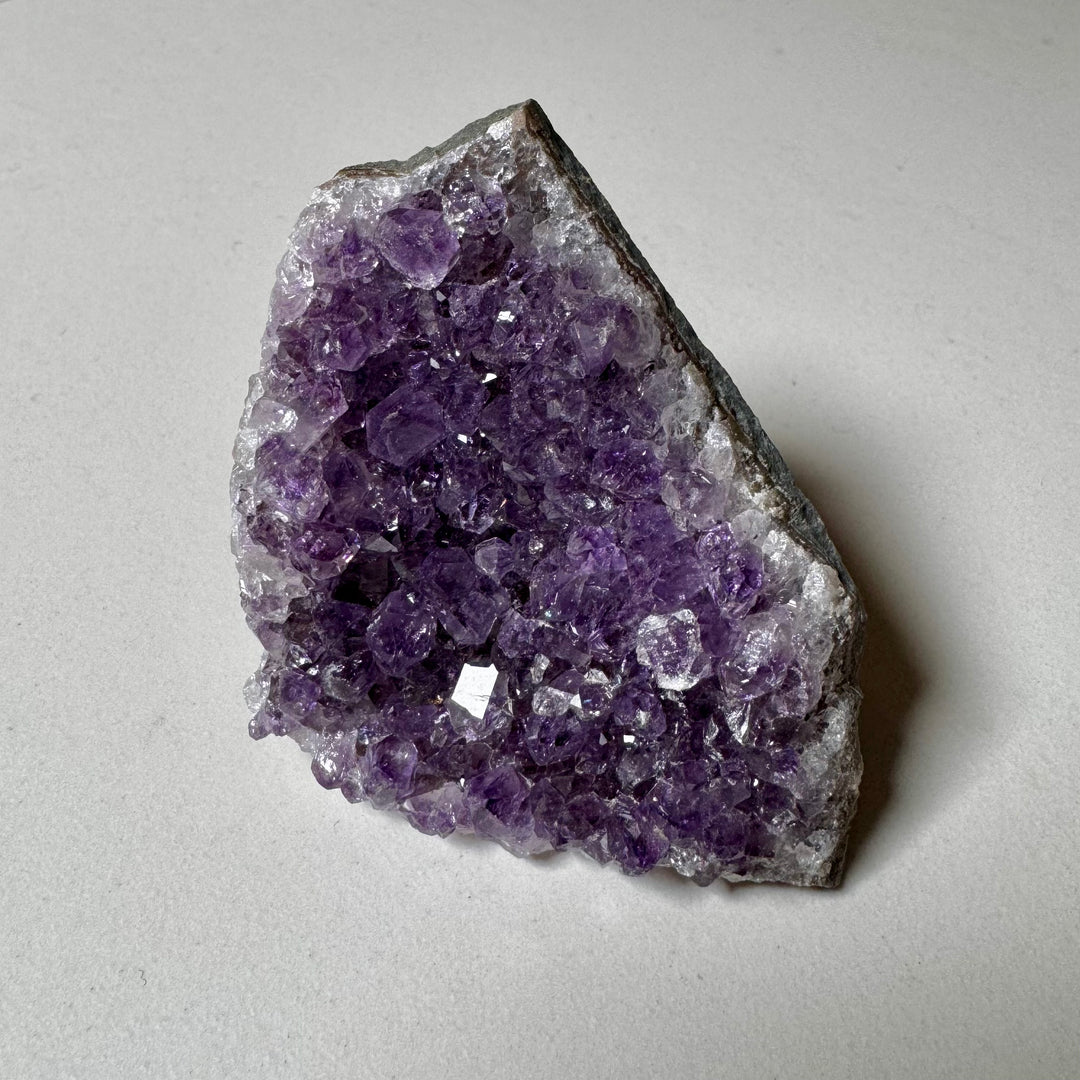 Ametistiklusterin pala yläviistosta kuvattuna; violetit kiteet kiinnityvät vihreänharmaaseen peruskiveen. - A piece of an amethyst cluster photographed from upper diagonal; purple crystals rising from a greenish-grey base rock.