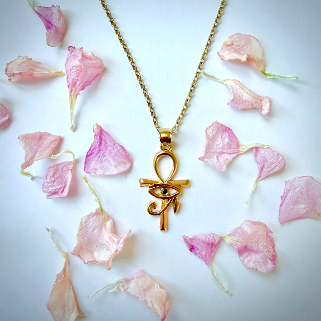 Lähikuva 18k kullatusta Ankh-risti- ja Horuksen silmä -riipuksesta valkoisella taustalla. Kultainen kaulakoru ja riipus, jonka keskellä on musta zirkonia, on ympäröity siroteltuilla vaaleanpunaisilla terälehdillä.