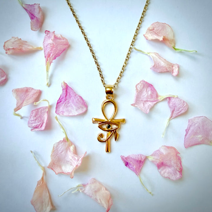 Lähikuva 18k kullatusta Ankh-risti- ja Horuksen silmä -riipuksesta valkoisella taustalla. Kultainen kaulakoru ja riipus, jonka keskellä on musta zirkonia, on ympäröity siroteltuilla vaaleanpunaisilla terälehdillä.