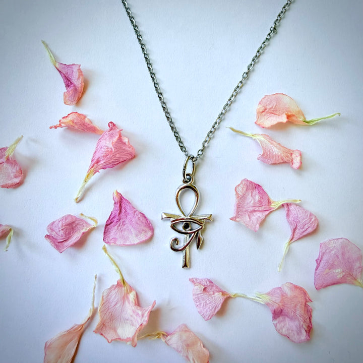 Yläviistosta otettu kuva hopeisesta Ankh-risti- ja Horuksen silmä -kaulakorusta valkoisella taustalla. Siro hopeaketju ja riipus, jossa on musta zirkonia-kivi, on aseteltu vaaleanpunaisten kukan terälehtien keskelle.