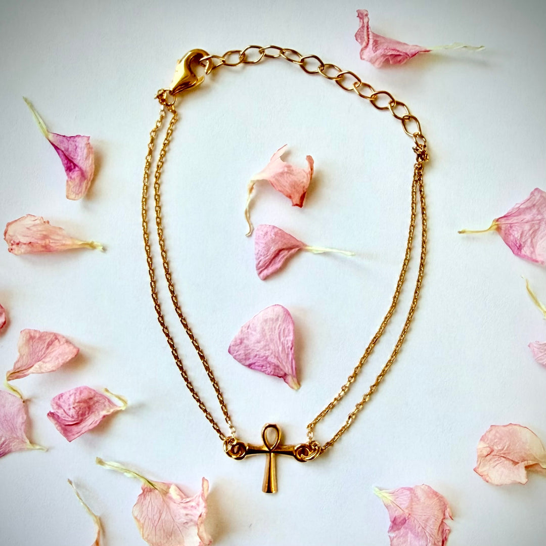 Kultainen rannekoru, jossa on pieni Ankh-risti -amuletti ja kaksinkertainen ketju, aseteltuna valkoiselle taustalle vaaleanpunaisten kukan terälehtien keskelle.