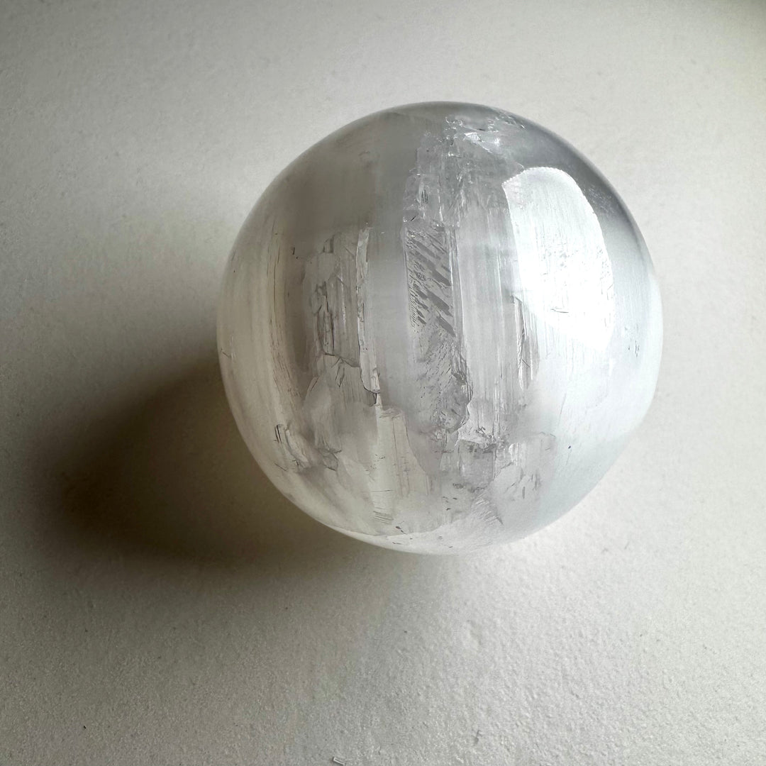 Kirkas ja läpikuultava seleniittipallo, jonka pinta heijastaa valoa silkkimäisesti. - Clear and translucent selenite sphere with a light-reflecting, silky surface.