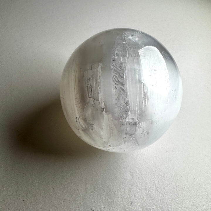 Kirkas ja läpikuultava seleniittipallo, jonka pinta heijastaa valoa silkkimäisesti. - Clear and translucent selenite sphere with a light-reflecting, silky surface.