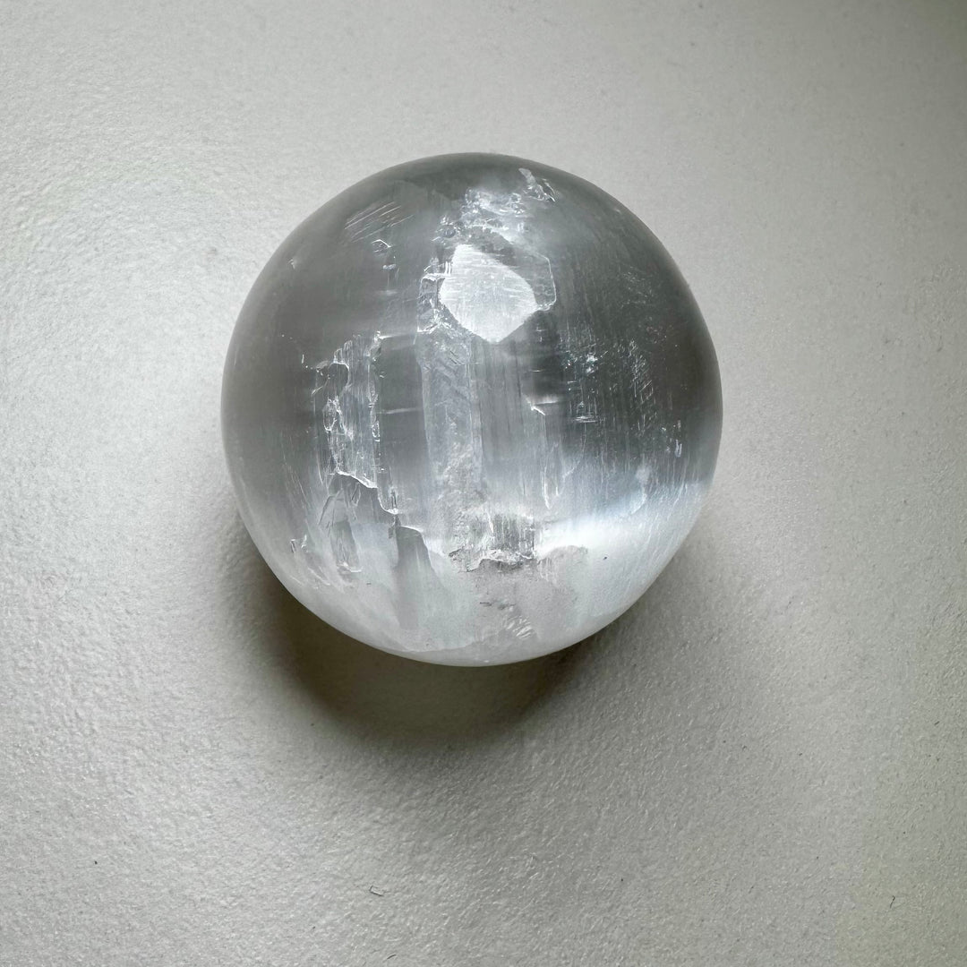 Kirkas ja läpikuultava seleniittipallo, jonka pinta heijastaa valoa silkkimäisesti. - Clear and translucent selenite sphere with a light-reflecting, silky surface.