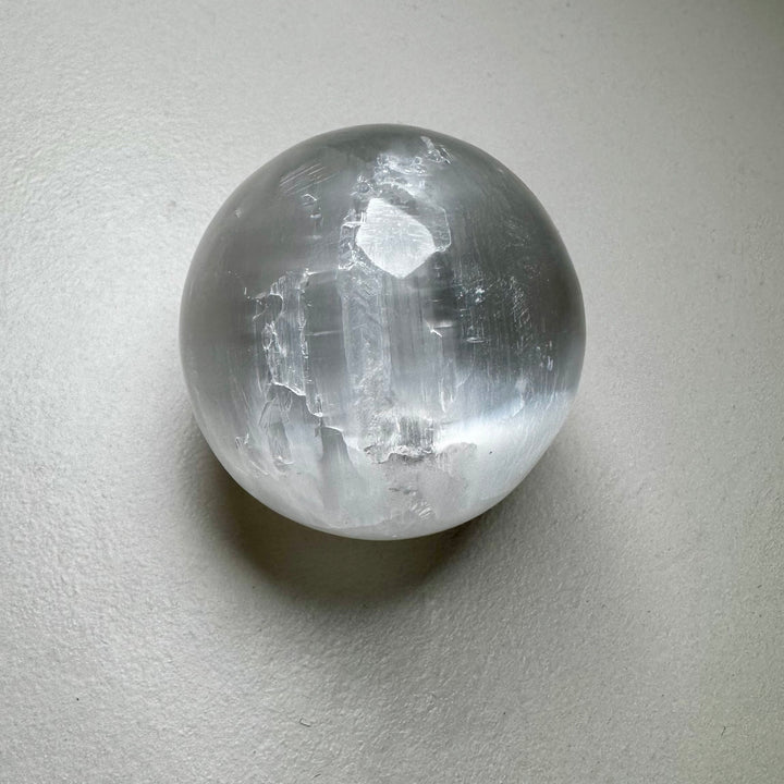 Kirkas ja läpikuultava seleniittipallo, jonka pinta heijastaa valoa silkkimäisesti. - Clear and translucent selenite sphere with a light-reflecting, silky surface.