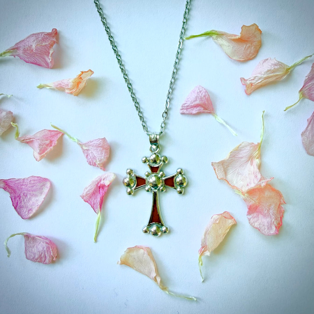 Hopeinen kaulakoru, jossa on punaisella emalilla täytetty kaldealainen risti -riipus. Ristin sakarat päättyvät pyöreisiin palloihin, ja keskellä on pyöreä kohokuvio. Risti on ketjussa ja makaa vaalealla alustalla, ympärillään irrallisia, kuivattuja vaaleanpunaisia ja kermanvärisiä ruusun terälehtiä.