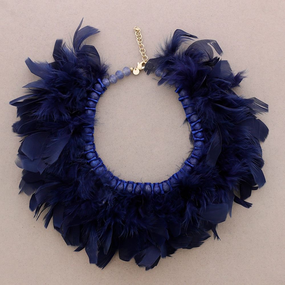 Syvän sininen sulkakaulakoru, jossa luonnonsulat on punottu helmillä koristeltuun nauhapohjaan. - Deep blue feather necklace with natural feathers woven into a beaded ribbon base.