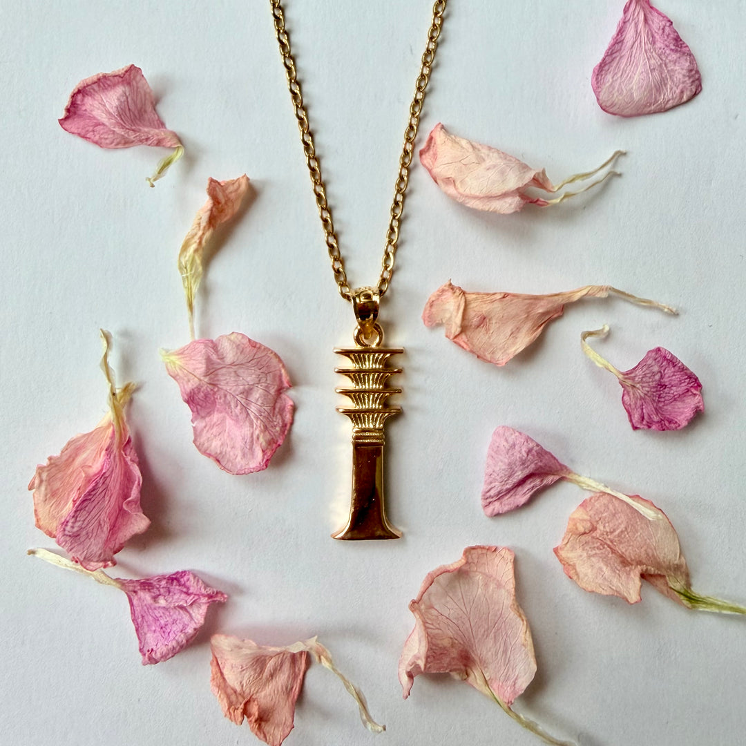 Kullattu djed-pilariamuletti ohuessa ketjussa, ympärillään vaaleanpunaisia kuivattuja kukan terälehtiä valkoisella taustalla. - A gilded djed pillar amulet on a thin chain, surrounded by pink dried flower petals on a white background.