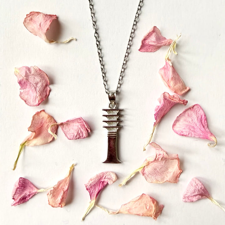 Hopeinen djed-pilariamuletti ketjussa, aseteltuna valkoiselle alustalle vaaleanpunaisten kuivattujen terälehtien keskelle. - A silver djed pillar amulet on a chain, placed on a white surface amidst pink dried flower petals.