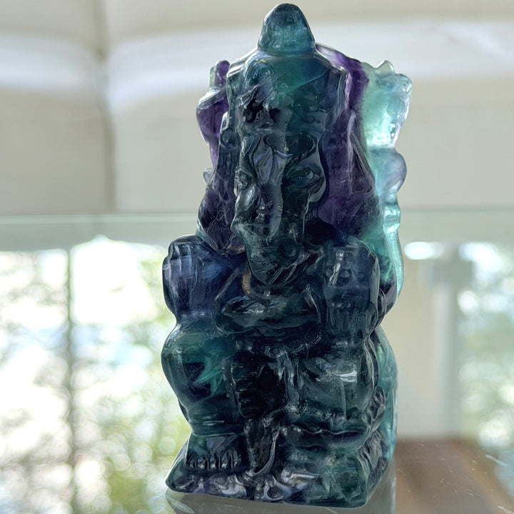 Fluoriitti Ganesha -figuuri sivulta kuvattuna, läpikuultava vihreänsininen ja violetti kivi korostuu valoa vasten.