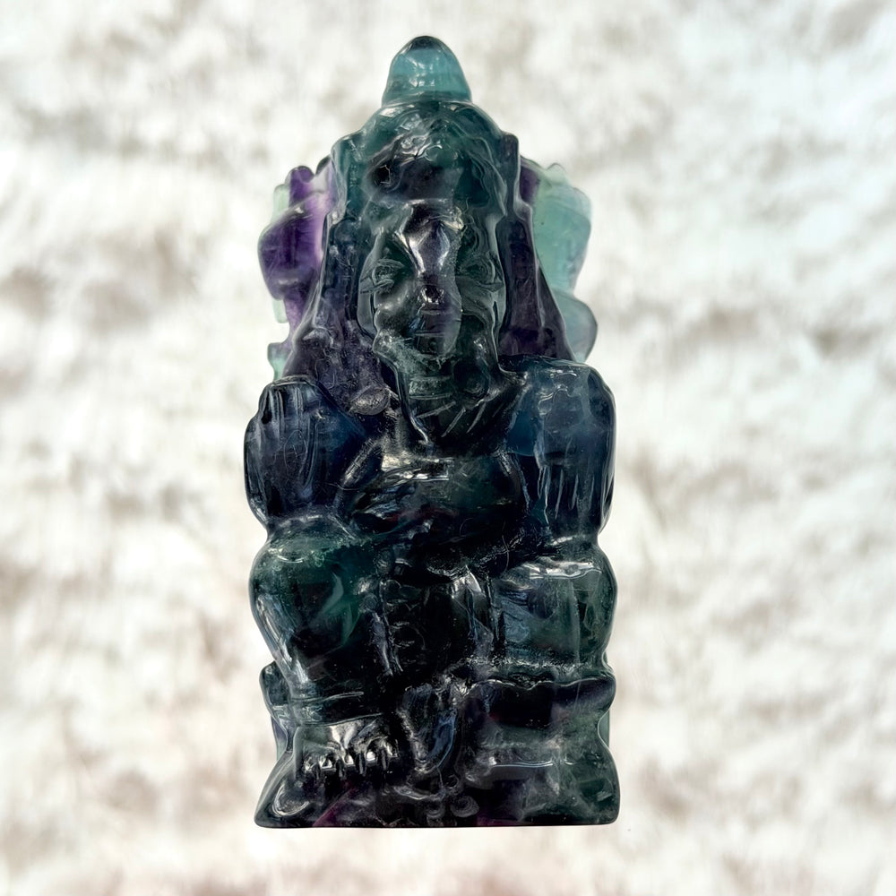 Fluoriitista veistetty Ganesha-patsas edestä kuvattuna, vihreän, sinisen ja violetin sävyinen läpikuultava kivifiguuri lasialustalla.