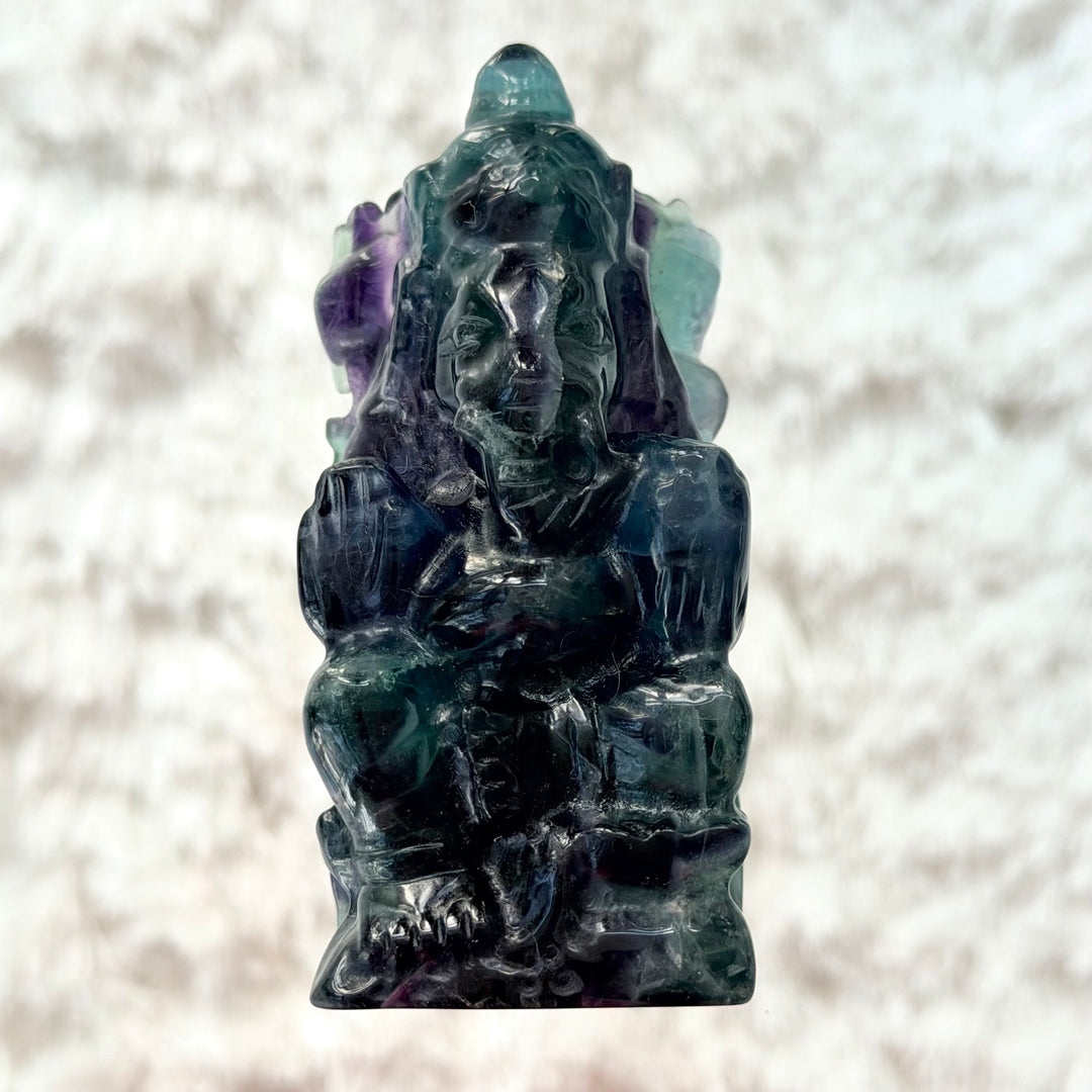 Fluoriitista veistetty Ganesha-patsas edestä kuvattuna, vihreän, sinisen ja violetin sävyinen läpikuultava kivifiguuri lasialustalla.