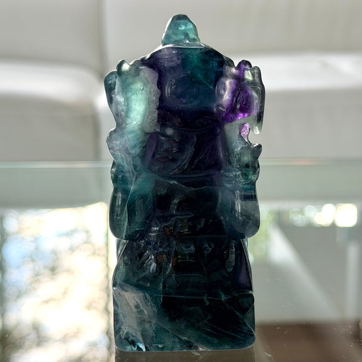 Fluoriitti Ganesha -patsaan takaosa, läpikuultava tumma kiviveistos violetilla ja turkoosilla sävyllä lasialustalla.