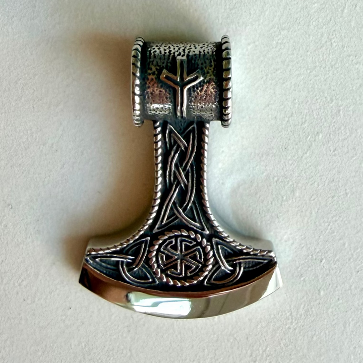 Hopeanvärinen Mjölnir-amuletti mustutetuilla punoskuvioilla ja geometrisilla koristeaiheilla vaalealla taustalla.