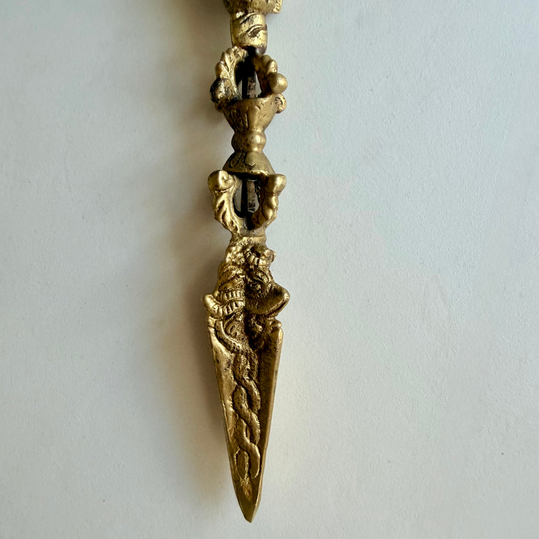 Lähikuva phurban kullanvärisestä terästä, kohokuvioisista käärmeaiheista ja kahvan alaosasta. / Close-up of the phurba’s gold-coloured blade, raised serpent motifs and lower handle.