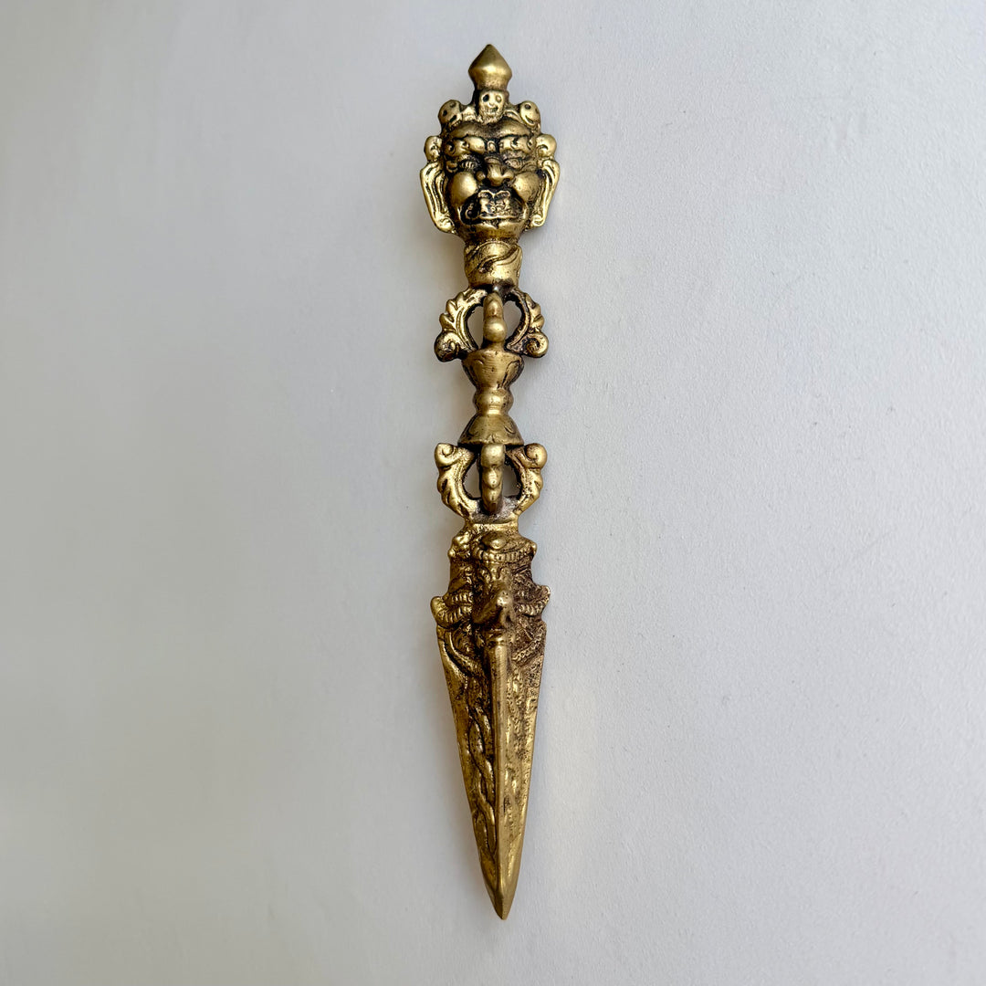 Kullanvärinen 21 cm phurba-rituaalitikari edestä, raivokas kasvoaihe, vajra-kahva ja kolmiteräinen terä. / Front view of a gold-coloured 21 cm phurba ritual dagger with a wrathful face, vajra handle and three-sided blade.