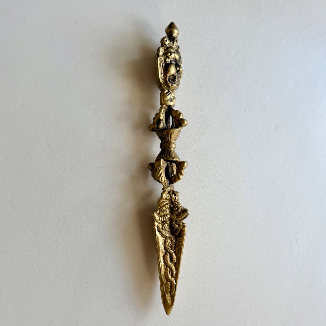 Kullanvärinen phurba viistosta kuvattuna, kasvoaihe, koristeellinen kahva ja terän kohokuviot. / Angled view of the gold-coloured phurba with a wrathful face, ornate handle and raised blade decoration.