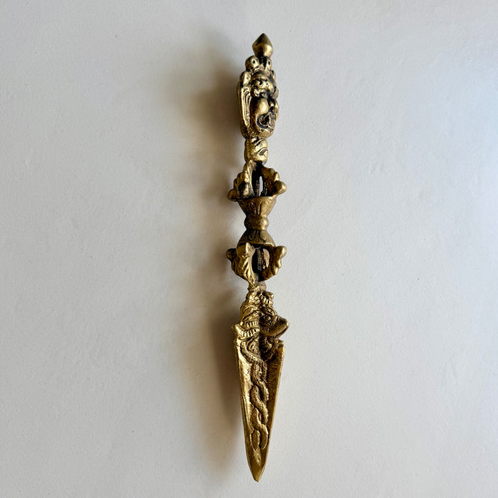 Kullanvärinen phurba viistosta kuvattuna, kasvoaihe, koristeellinen kahva ja terän kohokuviot. / Angled view of the gold-coloured phurba with a wrathful face, ornate handle and raised blade decoration.