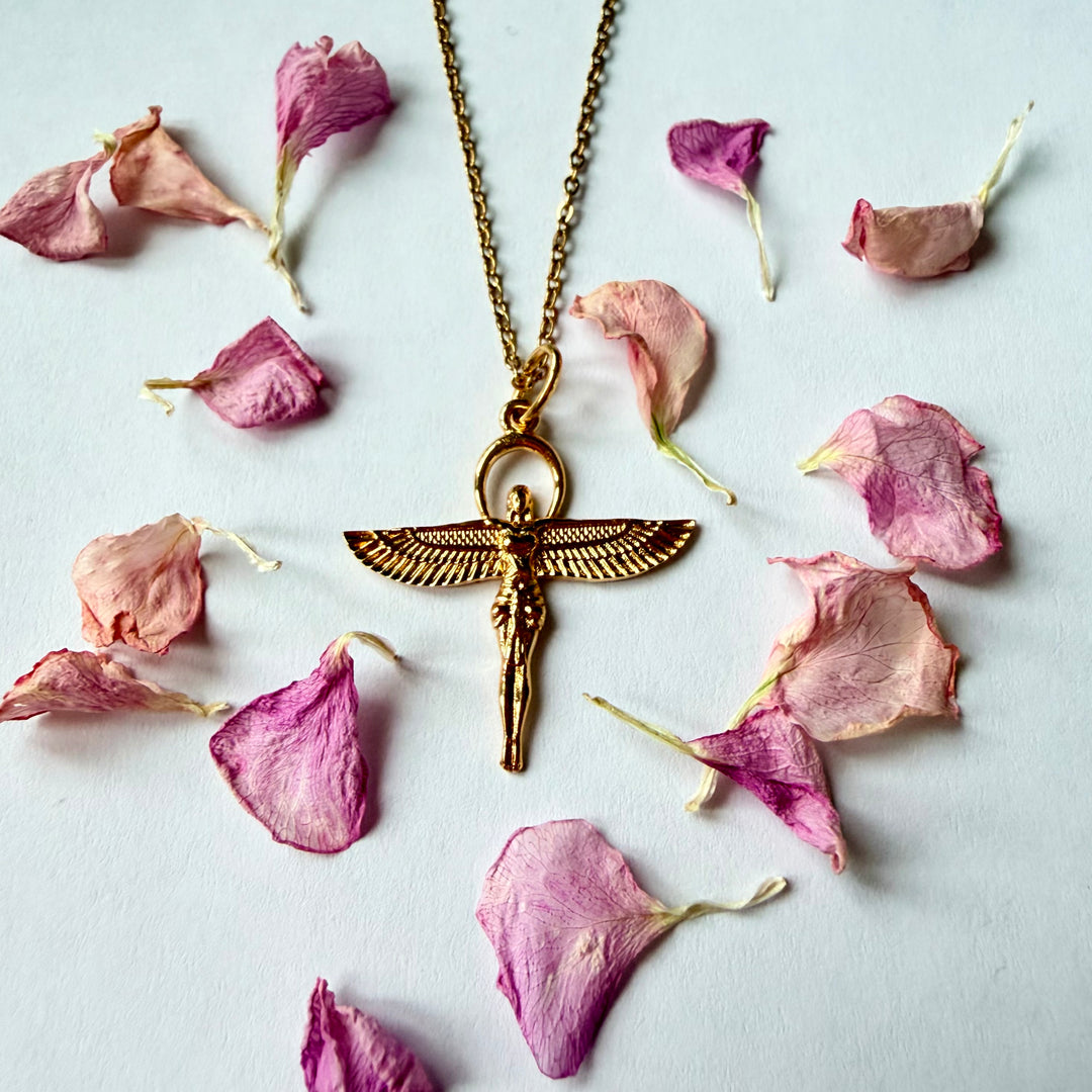 Lähikuva kultaisesta kaulakorusta, jossa on siivekäs Isiksen jumalatarhahmo. Koru lepää valkoisella pinnalla, ja sen ympärille on ripoteltu kauniisti vaaleanpunaisia ja purppuraisia kukan terälehtiä. - A close-up of a gold necklace with a winged Isis goddess pendant. The pendant lies on a white surface, framed by a delicate arrangement of pink and purple dried flower petals.