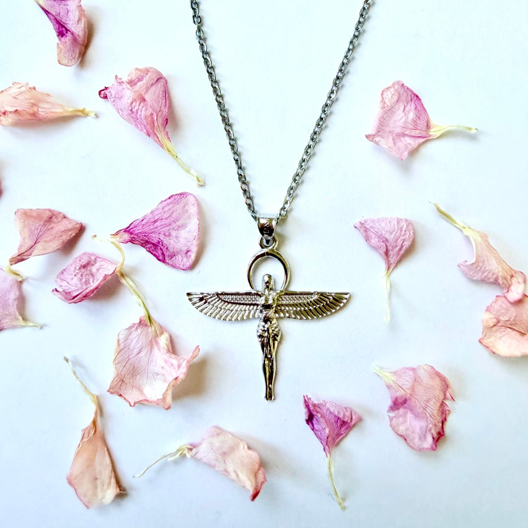 Yläviistosta otettu kuva hopeisesta Isiksen siivekästä jumalatarhahmoa esittävästä kaulakorusta. Koru on aseteltu valkoiselle taustalle, jota ympäröivät vaaleanpunaiset, kuivatut kukan terälehdet. - A top-down shot of a silver necklace featuring a winged goddess Isis pendant. The necklace is centered on a white background, artistically surrounded by scattered light pink dried flower petals.
