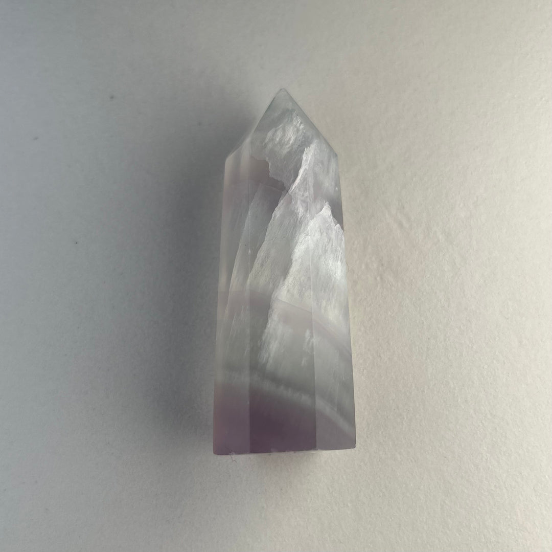 Vaalea laventelifluoriittikide sauvan muodossa, asetettuna vaalealle taustalle. - Light lavender fluorite crystal wand displayed on a pale background.
