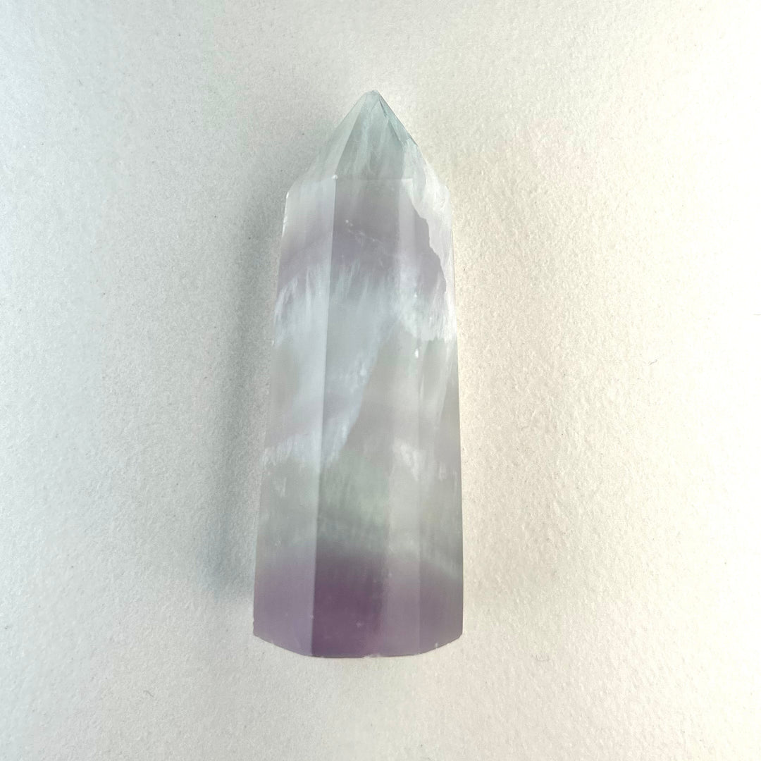 Puoliläpikuultava laventelifluoriittisauva, jonka pinnassa erottuu hienovaraisia violetteja sävyjä. - Semi-translucent lavender fluorite wand with delicate purple tones visible on its surface.