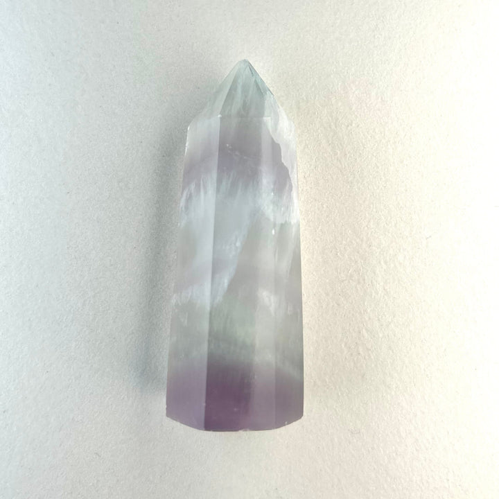 Puoliläpikuultava laventelifluoriittisauva, jonka pinnassa erottuu hienovaraisia violetteja sävyjä. - Semi-translucent lavender fluorite wand with delicate purple tones visible on its surface.