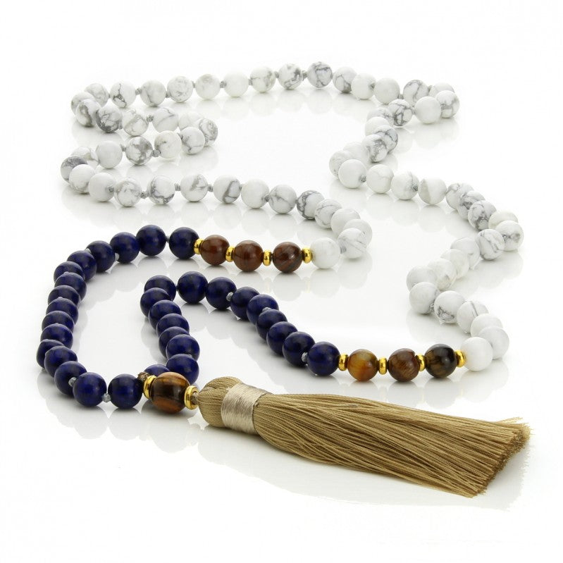 Malahelmet, jotka on valmistettu howliitista, sodaliitista ja tiikerinsilmästä, sekä beige tupsu nauhan päässä. - A mala made of howlite, sodalite, and tiger eye beads, featuring a beige tassel at the end.