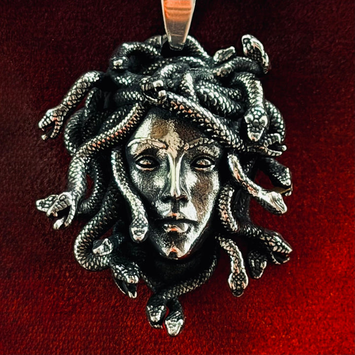 Medusa-amuletti punaisella taustalla, lähikuva tummasta patinoidusta hopeansävyisestä riipuksesta, jossa Medusan kasvot ja käärmehiukset.