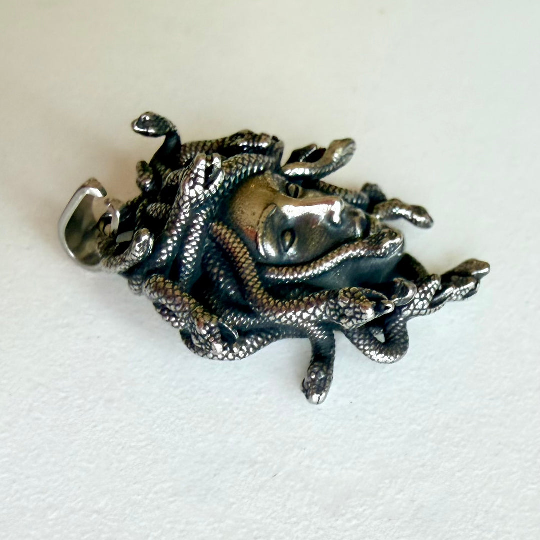 Medusa-riipus sivulta kuvattuna, kohokuvioinen käärmehiuksinen amuletti tummalla patinoidulla hopeansävyllä ja kiiltävällä pinnalla.