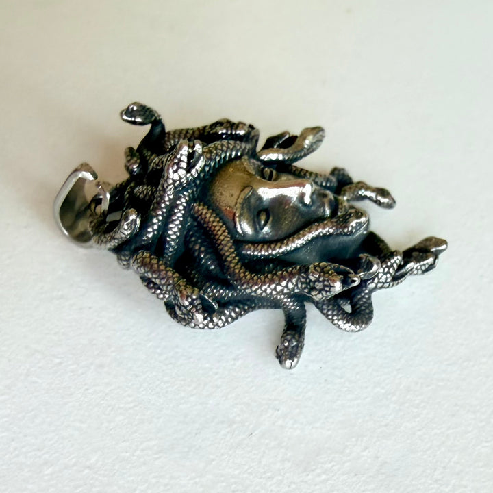 Medusa-riipus sivulta kuvattuna, kohokuvioinen käärmehiuksinen amuletti tummalla patinoidulla hopeansävyllä ja kiiltävällä pinnalla.