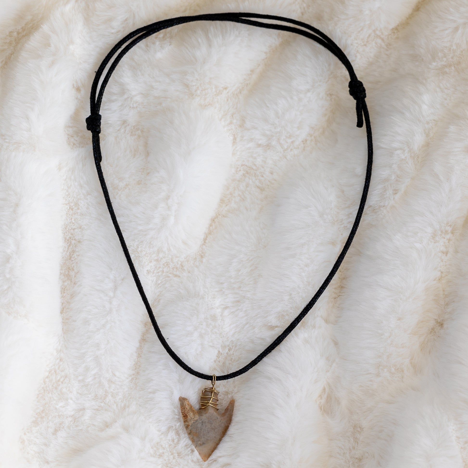 Koko kaulakoru, jossa musta tekonahkanyöri, piikivinen nuolenkärki ja kullanvärinen ripustuslenkki. - Full necklace with a black vegan leather cord, flint arrowhead, and gold-colored wire pendant loop.