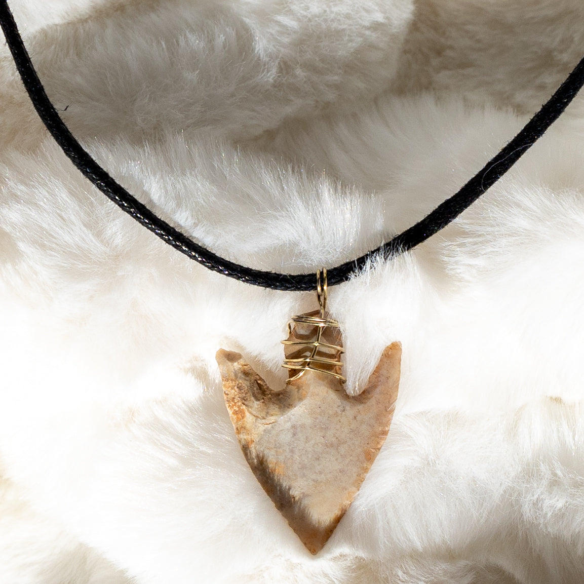 Lähikuva piikivisestä nuolenkärjestä kullanvärisellä metallilangalla ripustettuna mustaan tekonahkanyöriin. - Close-up of the flint arrowhead, attached with a gold-colored wire to a black vegan leather cord.