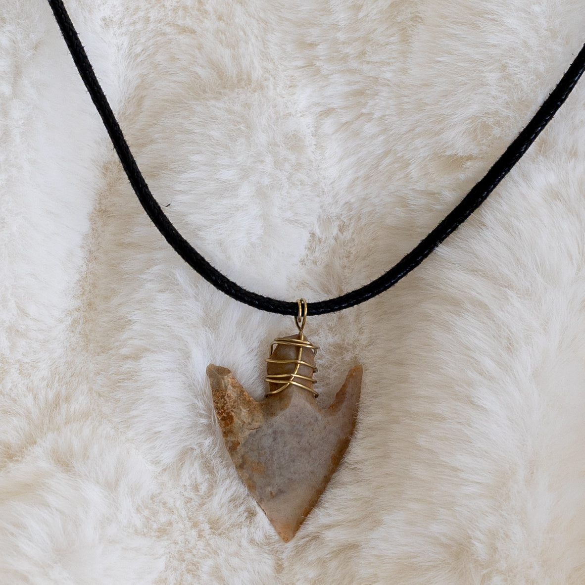 Lähikuva piikivisestä nuolenkärjestä kullanvärisellä metallilangalla ripustettuna mustaan tekonahkanyöriin.- Close-up of the flint arrowhead, attached with a gold-colored wire to a black vegan leather cord.