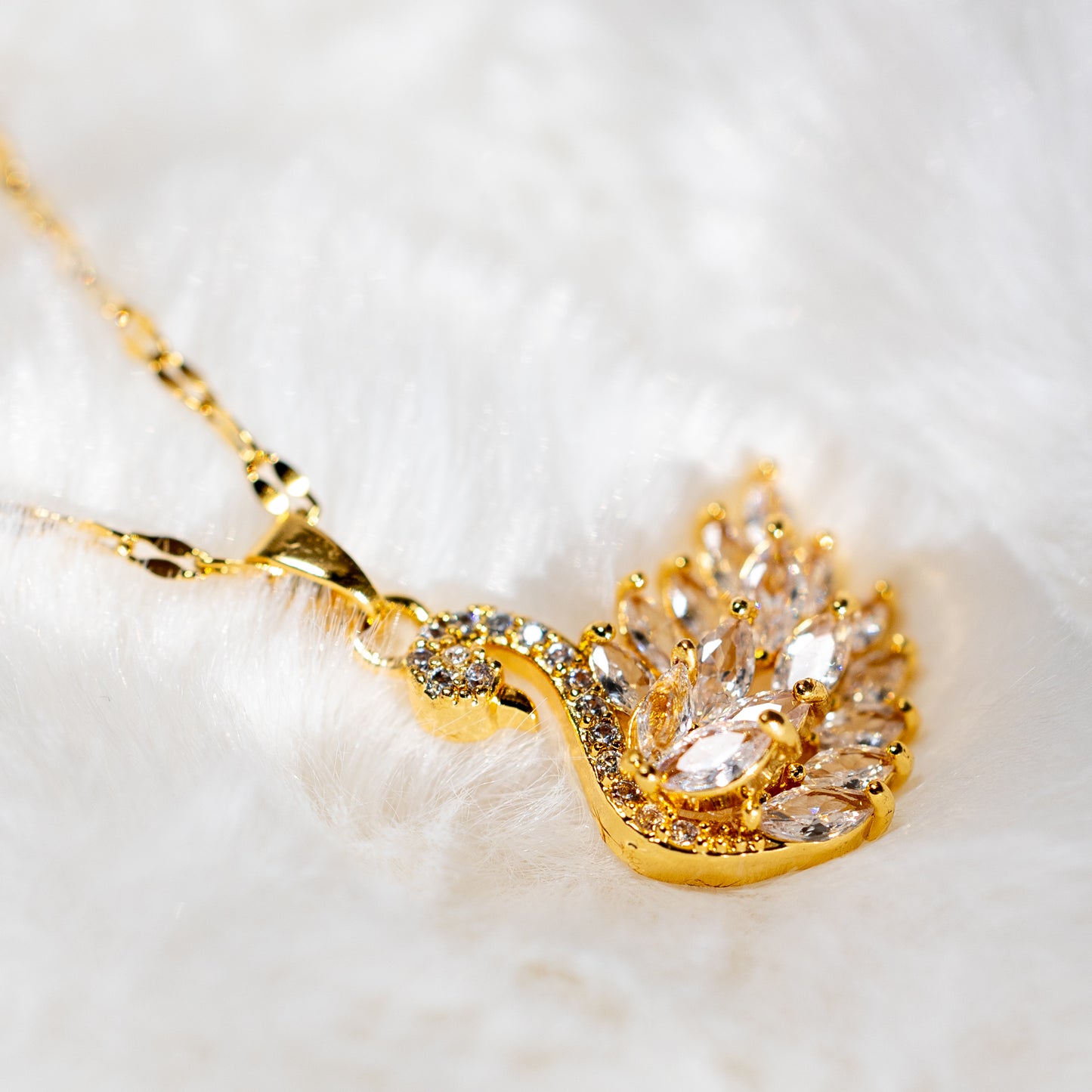 Lähikuva kullanvärisestä kaulakorusta ja sen joutsenamuletista, koristeltuna hiotuilla kivillä.  - A close-up photo of a gold-colored necklace featuring a stylish chain and a swan pendant adorned with faceted stones.