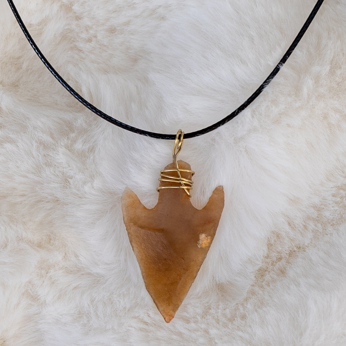  Lähikuva piikivisestä nuolenkärjestä kullanvärisellä metallilangalla ripustettuna mustaan tekonahkanyöriin. - Close-up of the flint arrowhead, attached with a gold-colored wire to a black vegan leather cord.