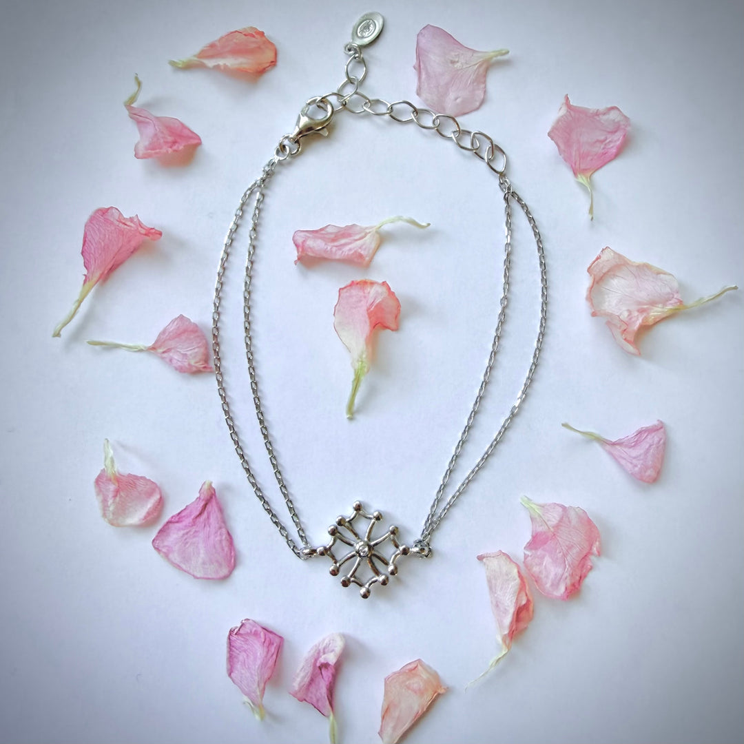 Yläviistosta otettu kuva hopeisesta rannerenkaasta, jossa on Oksitaaninen risti -amuletti. Kaksinkertainen ketju on aseteltu sydämen muotoon valkoiselle taustalle, jota ympäröivät vaaleanpunaiset kukan terälehdet. - A top-down shot of a silver bracelet featuring an Occitan cross charm. The double chain is arranged in a heart shape on a white background, framed by delicate pink flower petals.