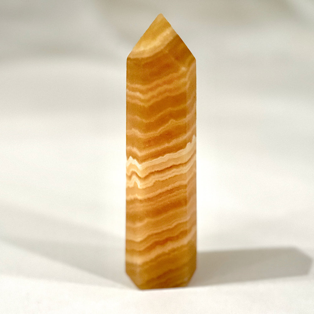 Hiottu oranssi kalsiittitorni, käytetty meditaatiossa ja energiakeskusten tasapainottamiseen, hehkuva ja kauniisti valoa heijastava. - Polished orange calcite tower, used for meditation and balancing energy centers, glowing and reflecting light beautifully.