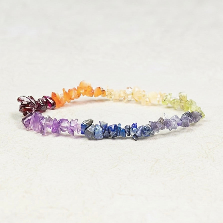 Chakra-rannerengas, jossa on värikkäitä hiottuja luonnonkivisiruja elastisessa nauhassa vaalealla taustalla. - Chakra bracelet with colourful polished natural stone chips on an elastic band against a light background.