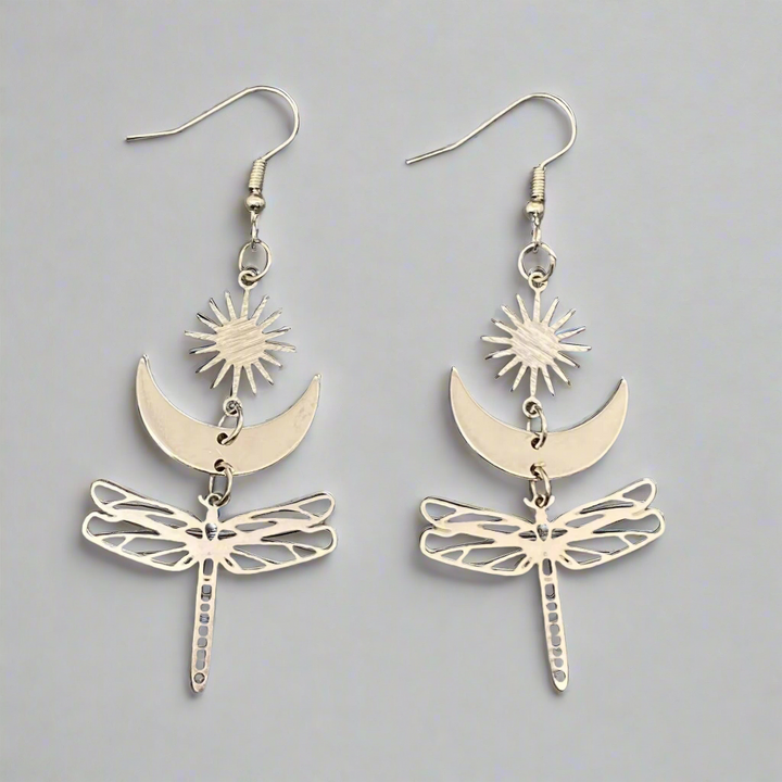 Hopeansävyiset kuparikorvakorut, joissa Aurinko, Kuu ja suuri sudenkorento symboloivat valoa, tunteiden rytmiä ja muodonmuutosta. - Silver-toned copper earrings featuring the Sun, the Moon, and a large dragonfly symbolizing light, the rhythm of emotions, and transformation.