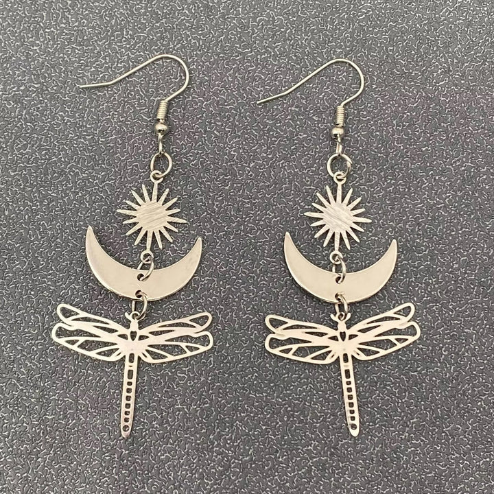Hopeansävyiset kuparikorvakorut, joissa Aurinko, Kuu ja suuri sudenkorento symboloivat valoa, tunteiden rytmiä ja muodonmuutosta. - Silver-toned copper earrings featuring the Sun, the Moon, and a large dragonfly symbolizing light, the rhythm of emotions, and transformation.