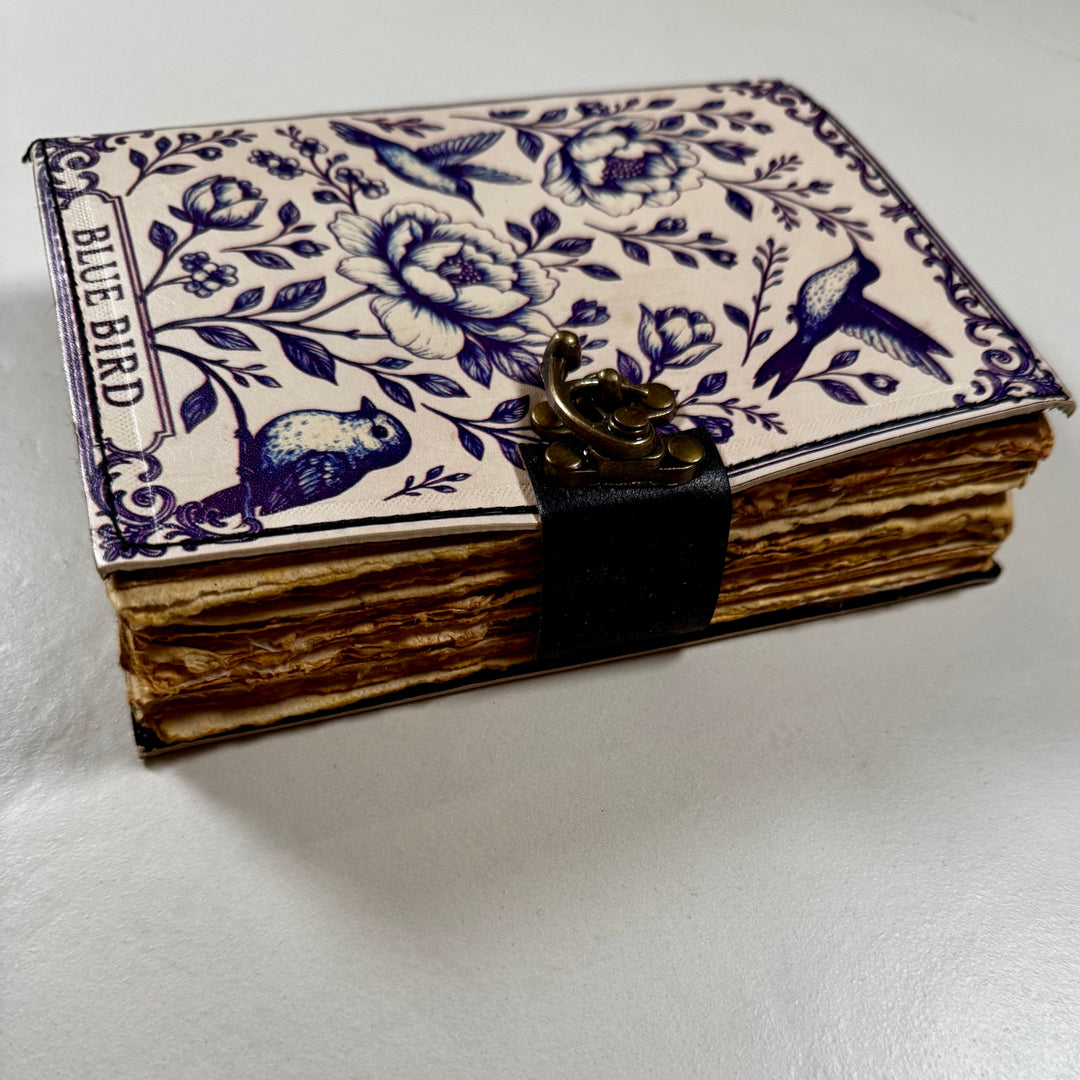 Sinilintuaiheinen nahkakantinen muistikirja sivusta kuvattuna, esillä kirjan paksuus ja lukko. - Blue bird-themed leather-bound notebook photographed from the side, showing the book’s thickness and lock.