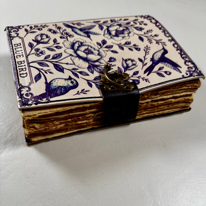 Sinilintuaiheinen nahkakantinen muistikirja sivusta kuvattuna, esillä kirjan paksuus ja lukko. - Blue bird-themed leather-bound notebook photographed from the side, showing the book’s thickness and lock.