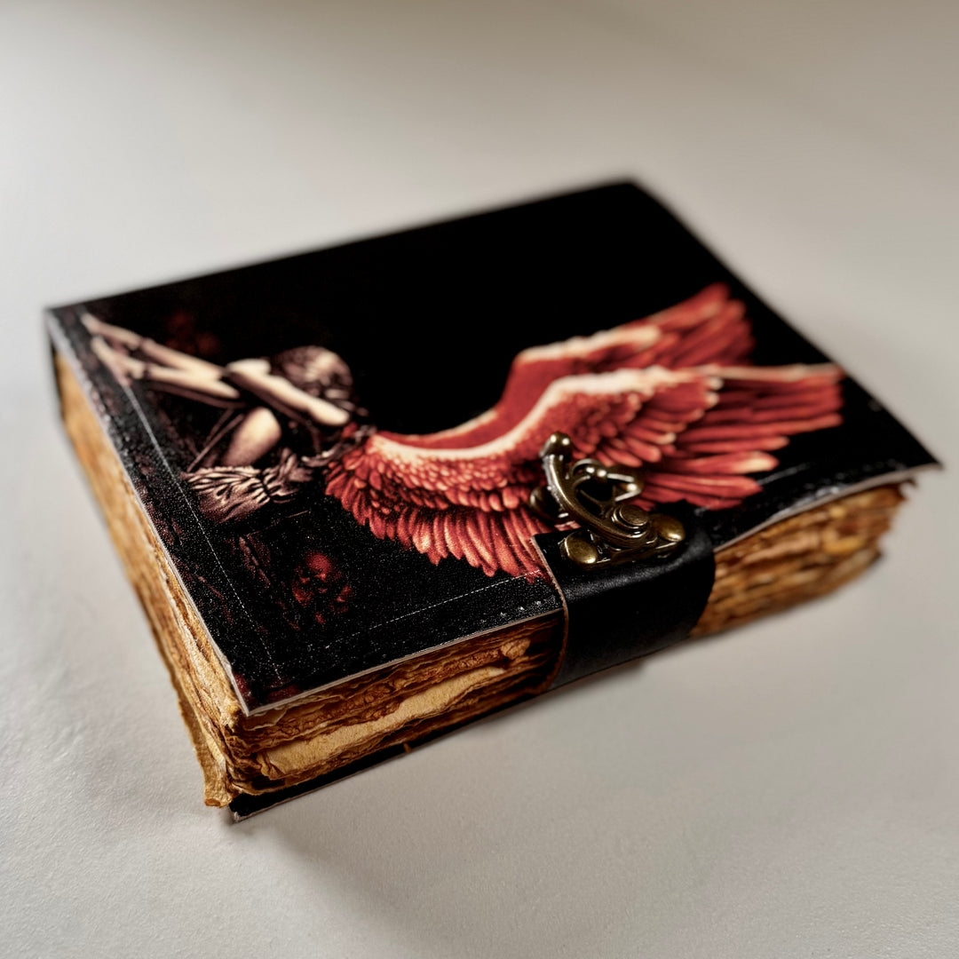 Enkeliaiheinen nahkakantinen muistikirja sivusta kuvattuna, esillä kirjan paksuus ja lukko. - Broken Angel -themed leather-bound notebook photographed from the side, showing the book’s thickness and lock.