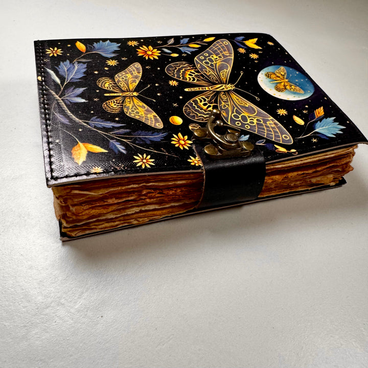 Perhosaiheinen nahkakantinen muistikirja sivusta kuvattuna, esillä kirjan paksuus ja lukko. - Golden Butterfly -themed leather-bound notebook photographed from the side, showing the book’s thickness and lock.