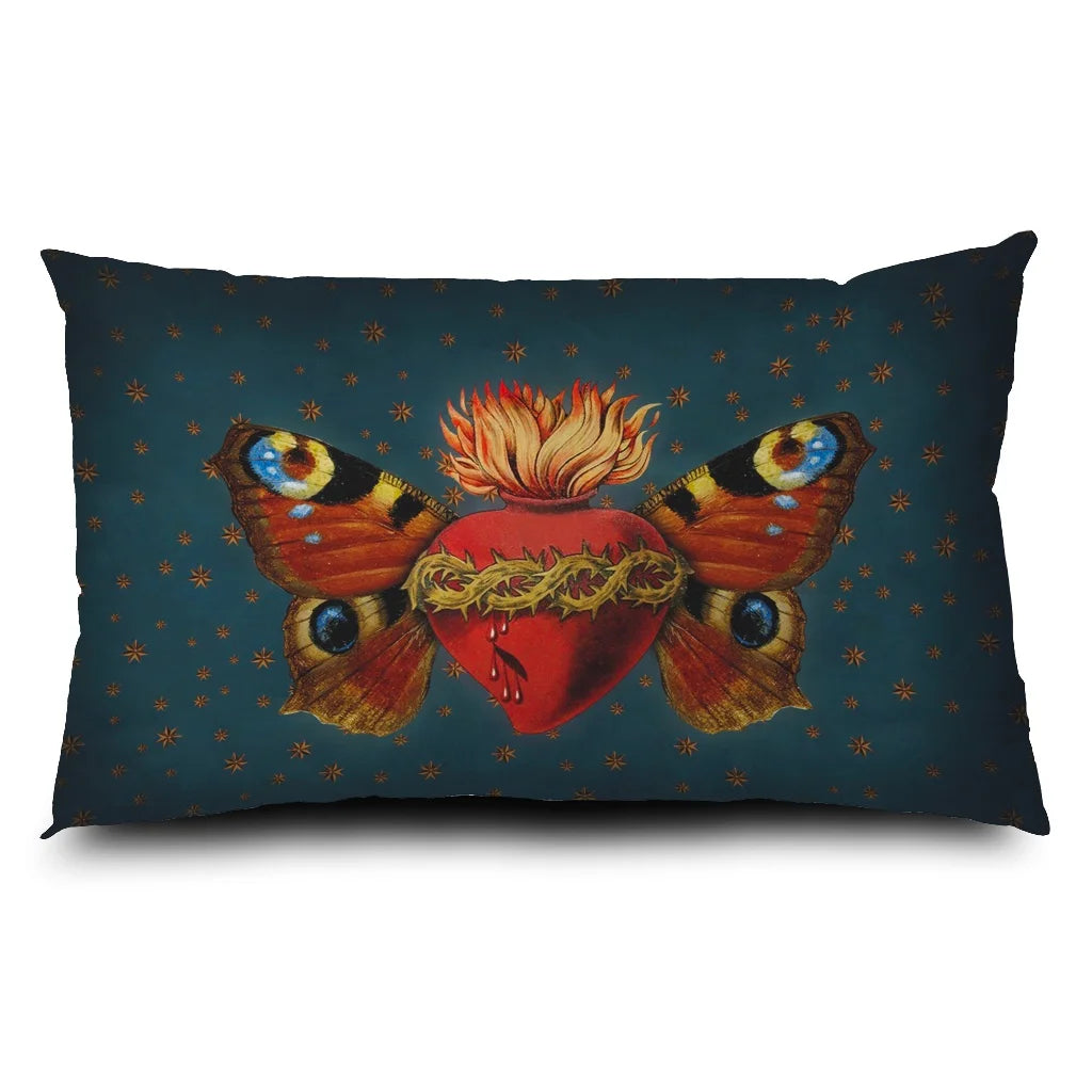 Kuvassa Voglio Bene -tyyny Sacred Butterfly -kuvituksella. Sydän, jota puristavat orjantappurat, liekehtii yläosastaan ja sillä on perhosen siivet. Taustalla sinisellä pohjalla tähtikuvioita.
The Voglio Bene cushion with Sacred Butterfly illustration. A heart encircled by thorns emits flames from its top and has butterfly wings. The blue background has starry ornaments.