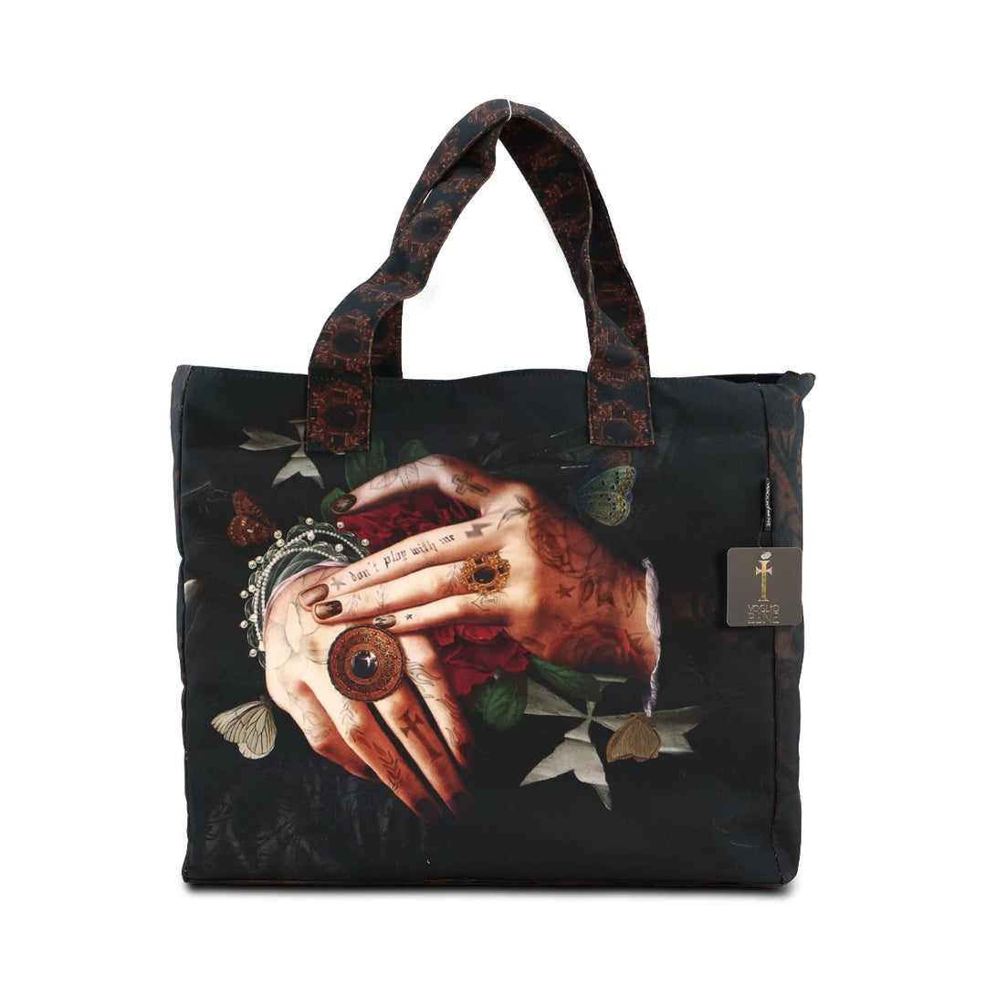 Kuvassa Voglio Bene -tote bag Protection -kuvituksella. Tatuoidut kädet lepäävät limittäin, sormissa tummia sormuksia ja käsien alla punainen kukka, taustana tumma goottinen kuviointi.
The Voglio Bene tote bag with Protection illustration. Tattooed hands rest intertwined, adorned with dark rings, with a red flower beneath them against a dark gothic background.